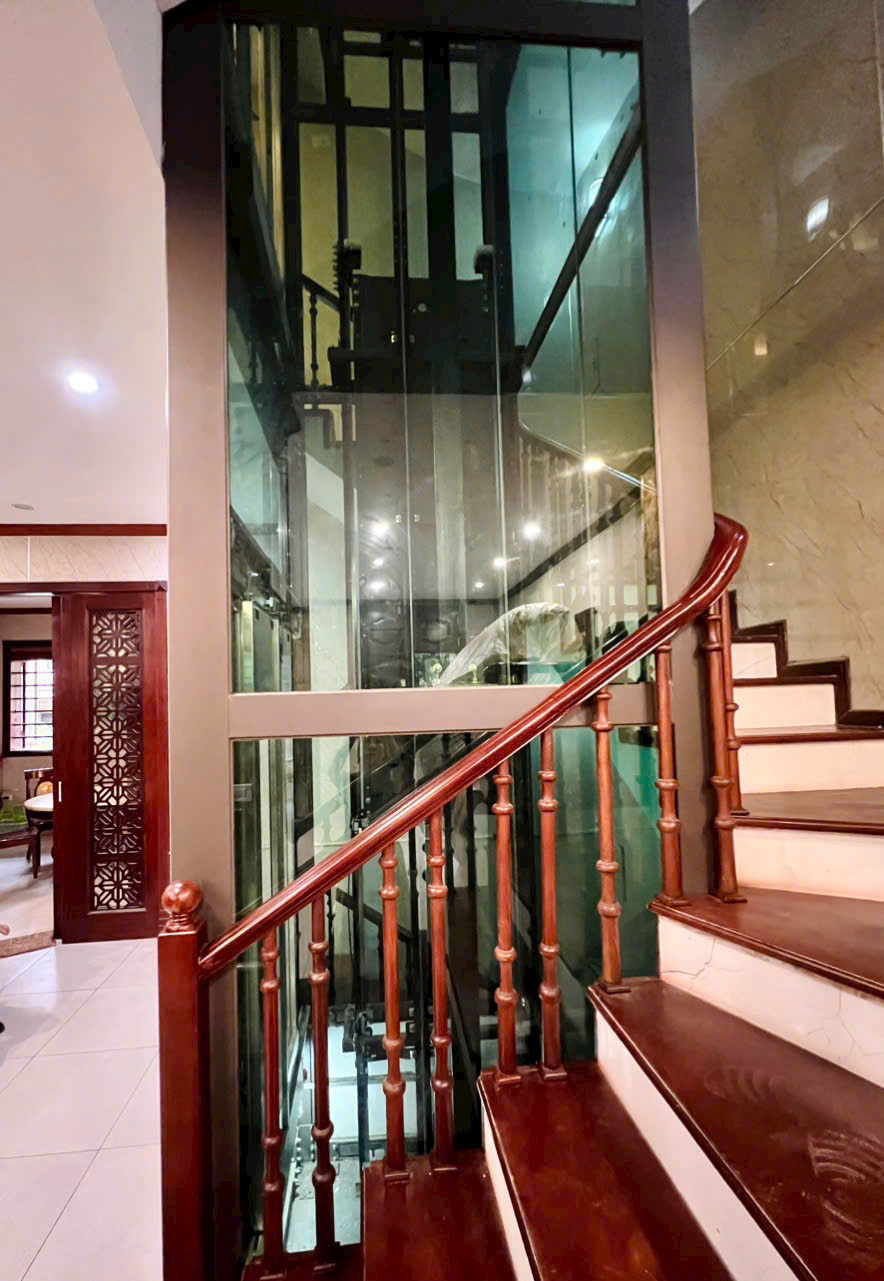 Thang máy gia đình vách kính đặt cạnh cầu thang gỗ trong nhà ở cao tầng